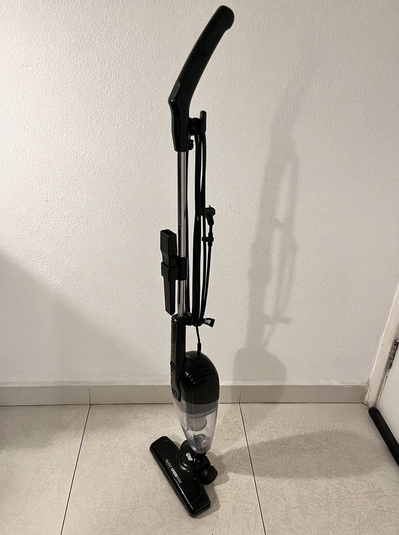 Aspirador vertical WAP preto, de corpo inteiro, apoiado numa parede branca, com o cabo enrolado no tubo.