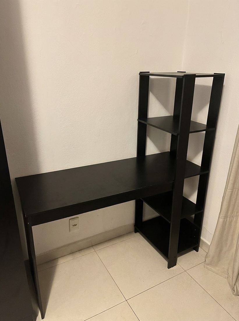 Escrivaninha preta com estante acoplada de quatro nichos, vazia, encostada na parede do quarto.