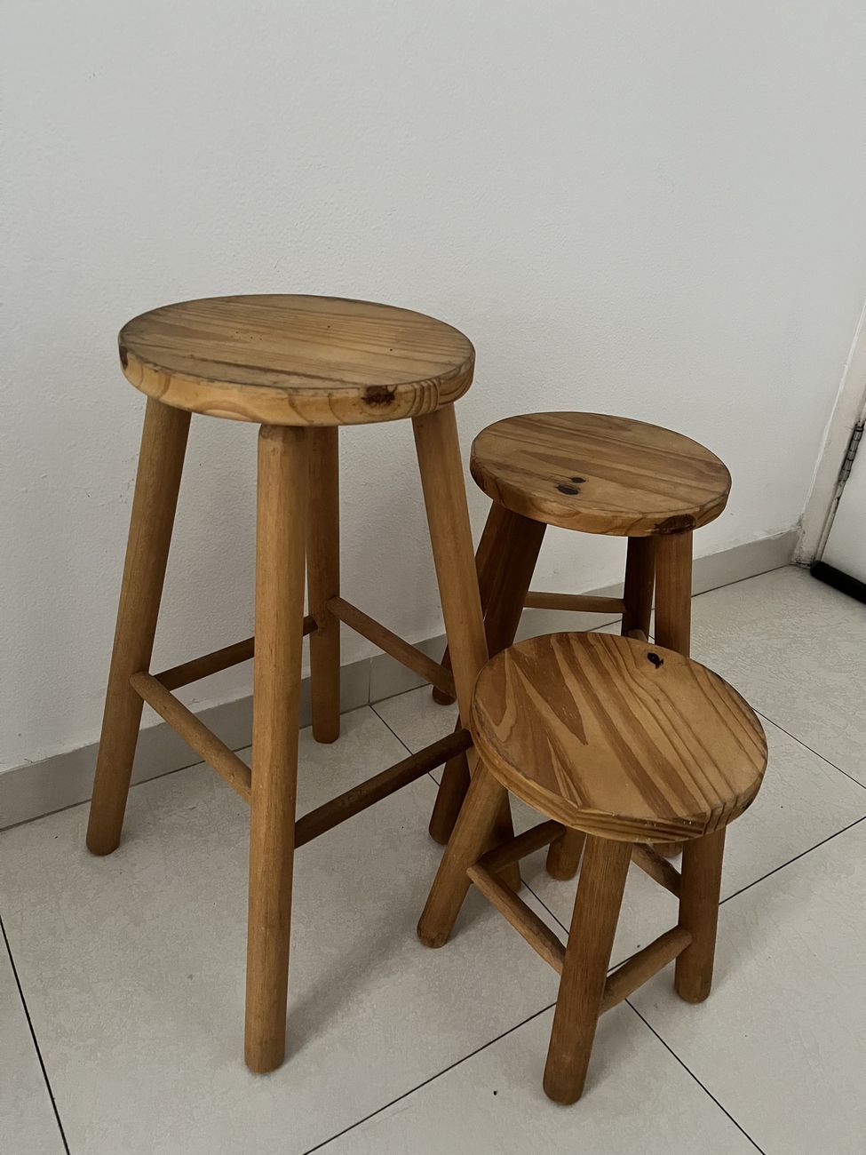 Três banquetas de madeira de alturas diferentes, agrupadas junto a uma parede branca.