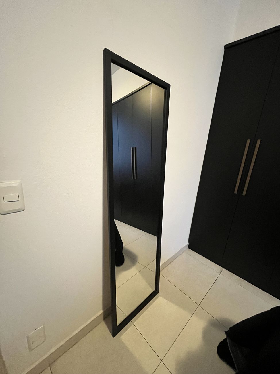 Espelho de corpo inteiro com moldura preta fina, encostado na parede ao lado de um guarda-roupa preto.