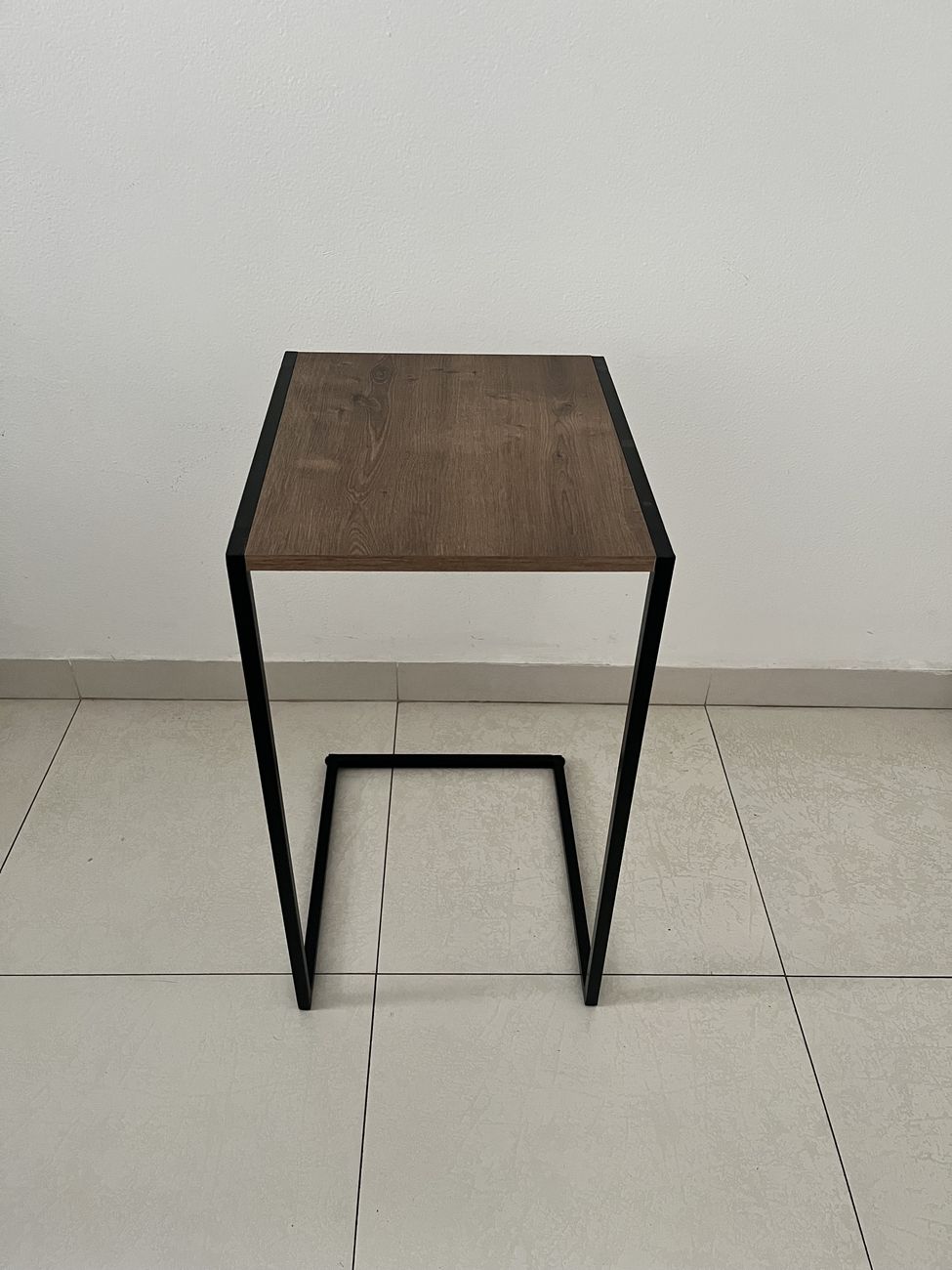 Mesa lateral em formato de C, com tampo amadeirado e estrutura de metal preto, vista de frente.