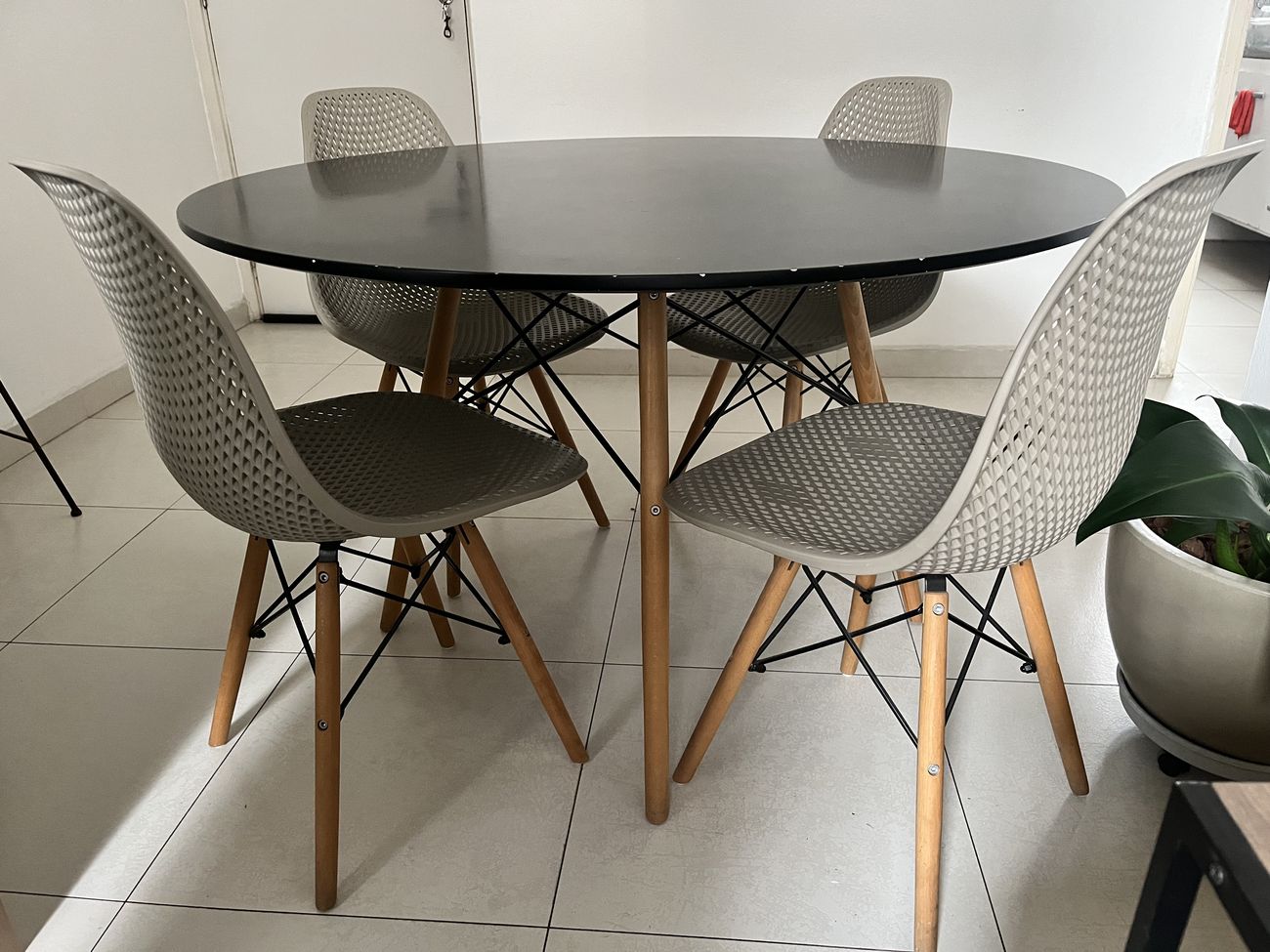 Mesa de jantar redonda com tampo preto e pés de madeira clara, com quatro cadeiras bege de encosto perfurado ao redor.