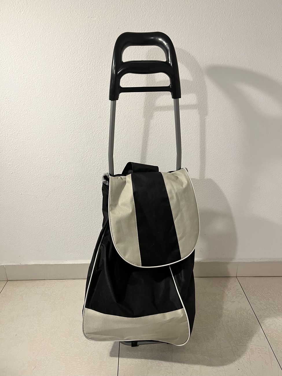 Carrinho de feira de duas rodas com bolsa preta e bege, visto de frente, encostado numa parede branca.