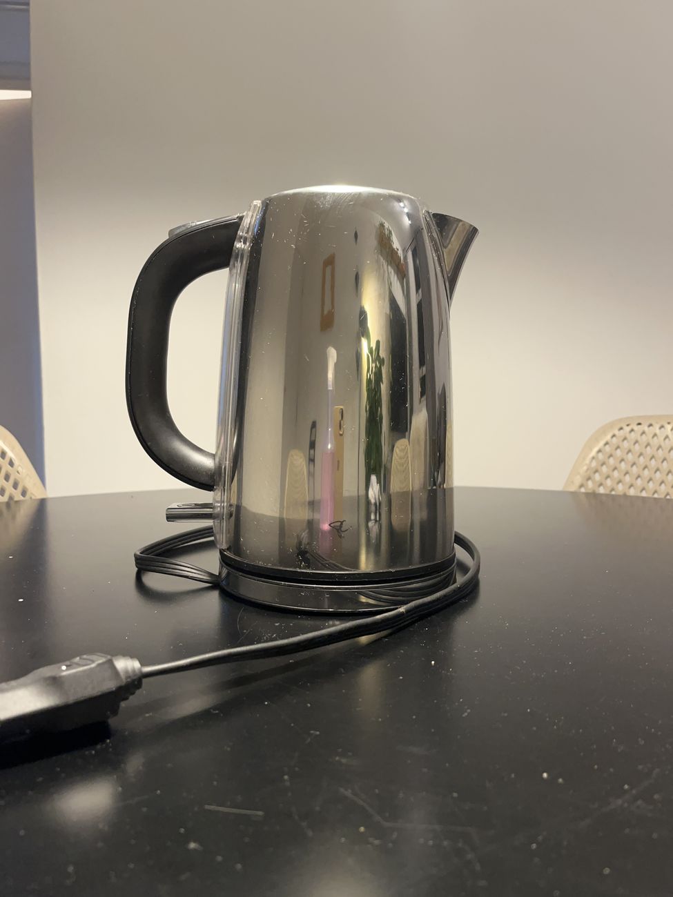 Chaleira elétrica de aço inox polido com alça preta, vista de frente sobre uma mesa preta.
