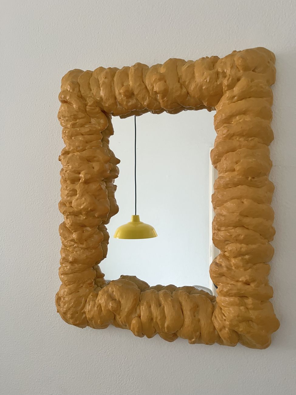 Espelho retangular com moldura escultural em relevo, cor mostarda, pendurado numa parede branca, refletindo uma luminária amarela.